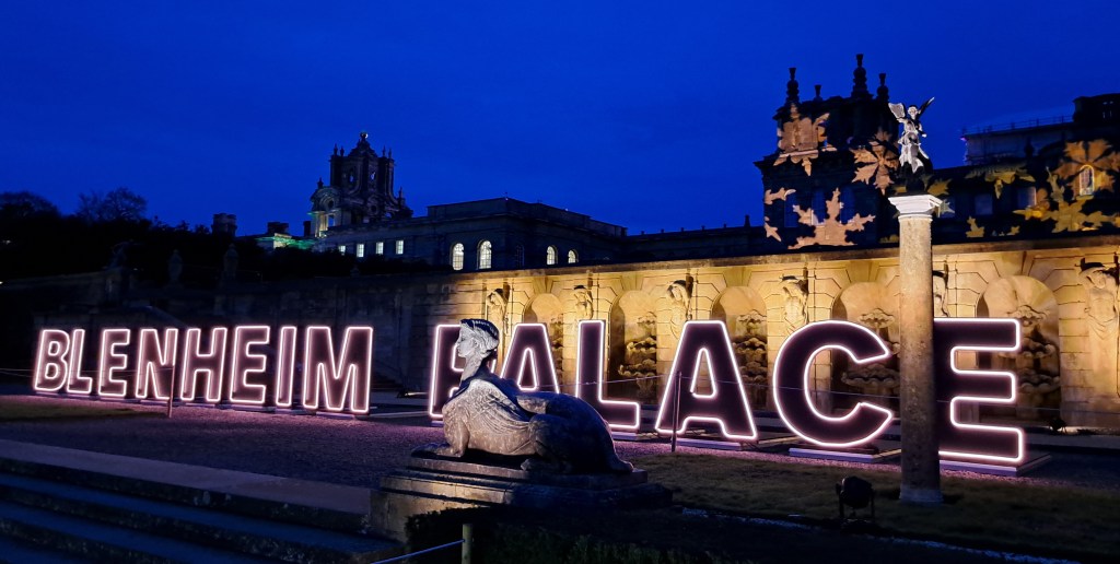 Illuminated sign for Blenheim Palace glowing in the evening light against the historical building, with a dark blue sky in the background.