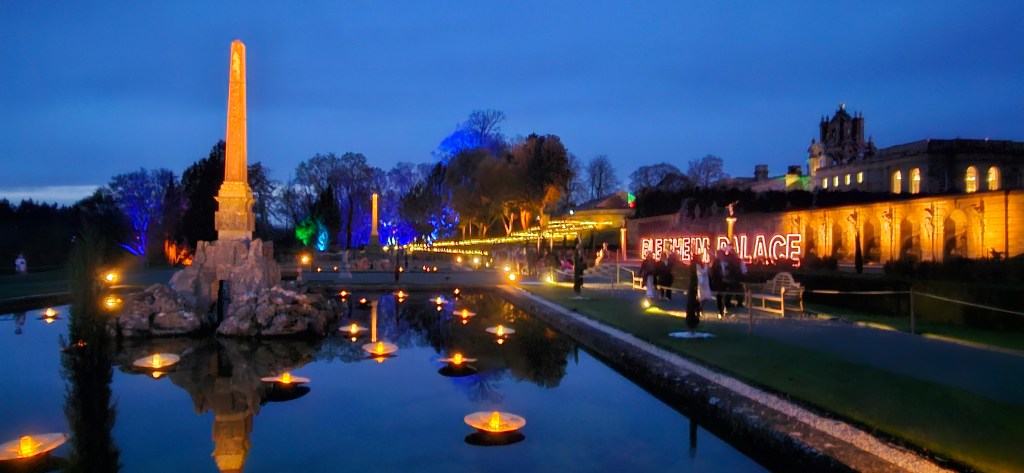 A nighttime view of Blenheim Palace illuminated with colorful lights, reflecting in a pond with floating lanterns, during the Christmas festivities.