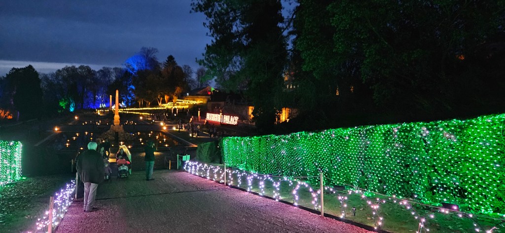 View of Blenheim Palace illuminated with colorful lights, with visitors walking along a pathway lined with illuminated greenery and a fountain in the foreground.