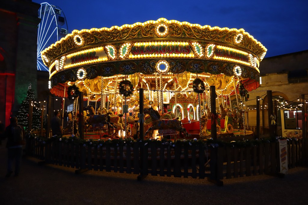 A beautifully illuminated vintage carousel surrounded by festive decorations at Blenheim Palace, with a Ferris wheel in the background and a twilight sky.